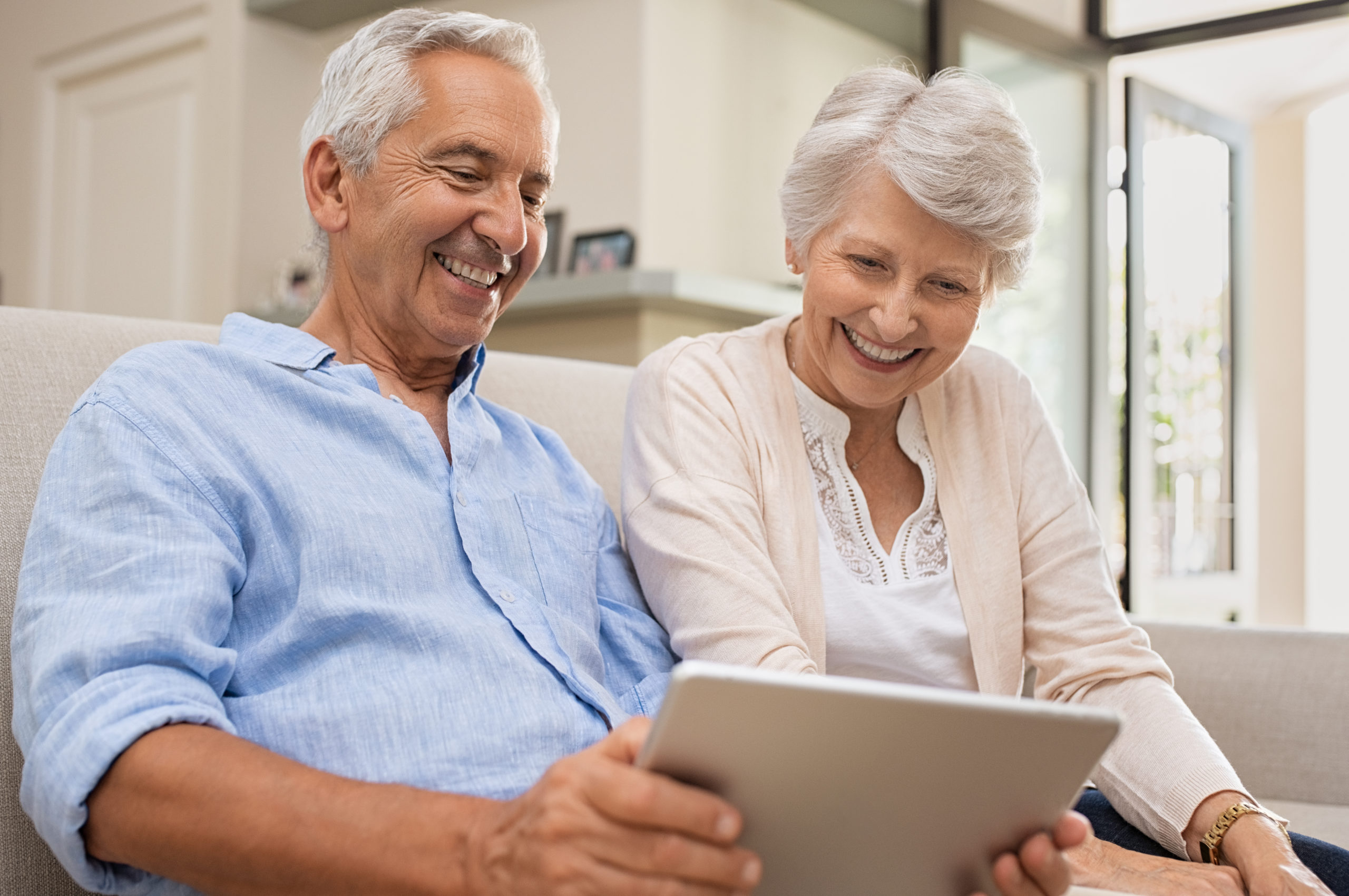 Senior couple using digital tablet sitting on couch at home. Happy old man surfing the net with his wife. Retired man and smiling elderly woman planning on summer vacation using tablet.